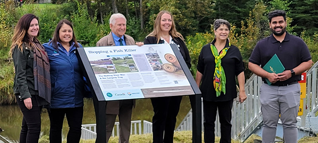 In the foreground, six people are standing around and behind a sign that is installed in the ground. The sign has pictures and information about sea lampreys and sea lamprey traps. A river is in the background as well as metal steps leading down to a platform at the water’s edge, which houses a sea lamprey trap.